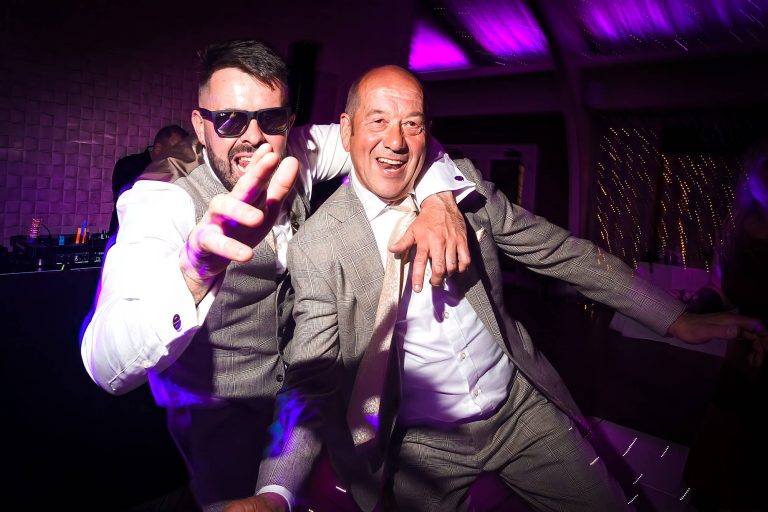 Groom and guest dancing in sunglasses under purple lights