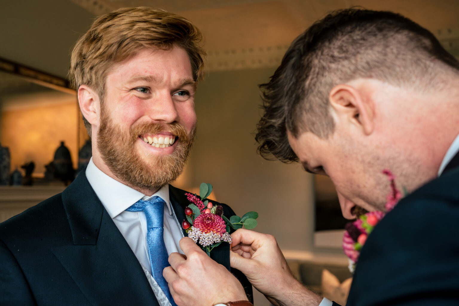Groom laughing as his best mate pins on his buttonhole during groom prep