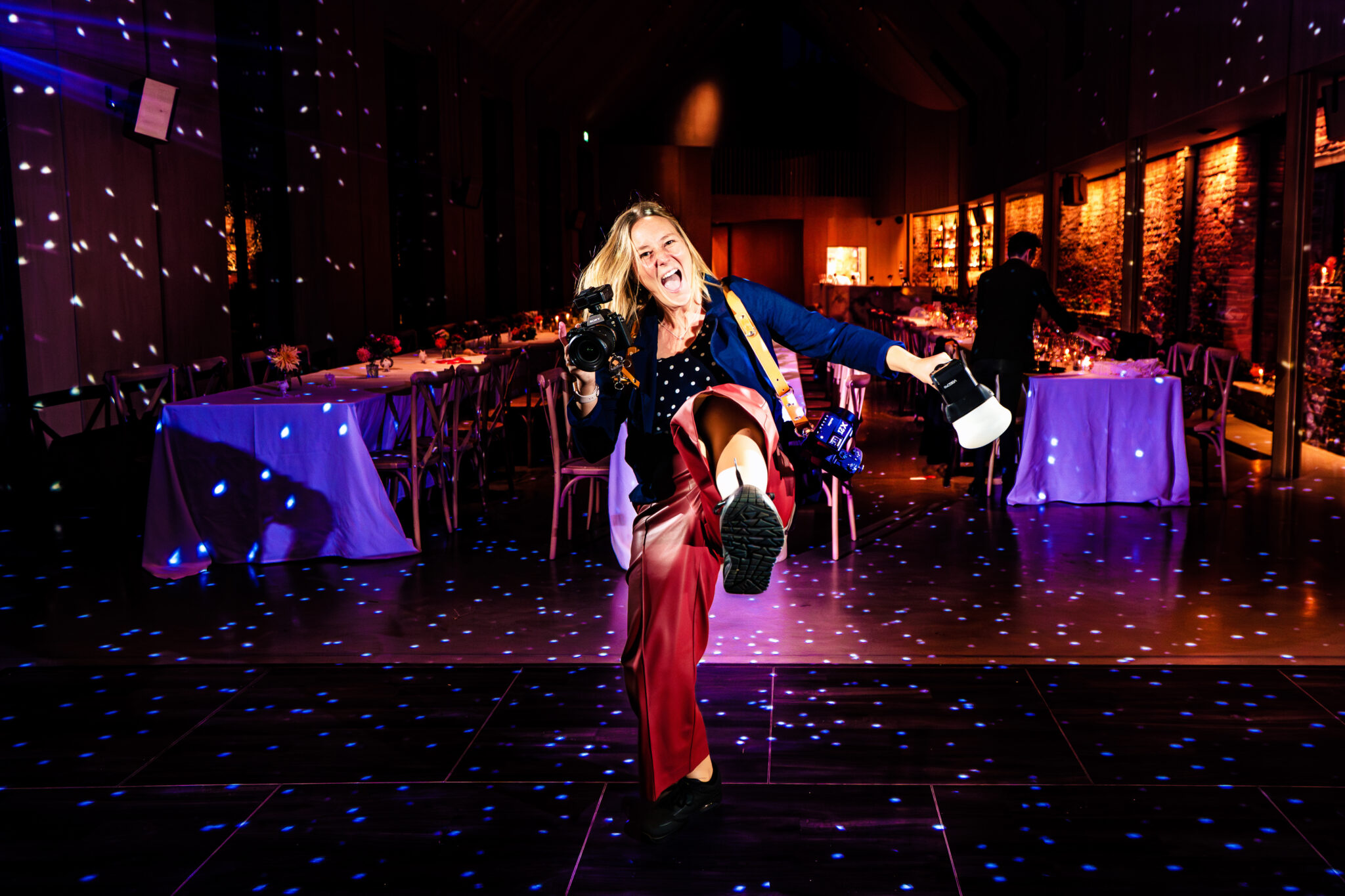 Emma Staley kung fu kicking the air on the dancefloor with her camera in one hand and a flash on a stick in the other