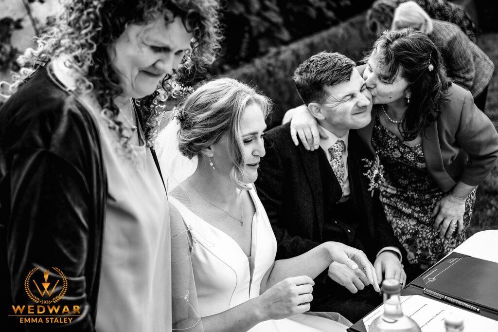 Bride and groom signing the register surrounded by family, black and white, WedWar 2026 award winning