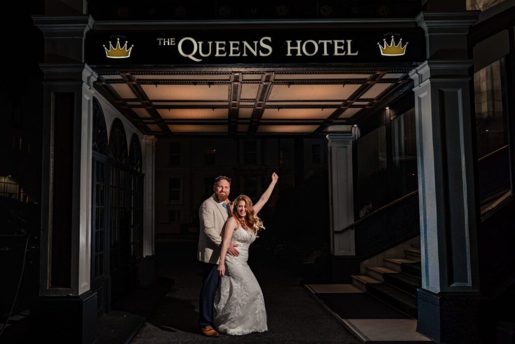 Bride throwing her arm in the air celebrating with the groom under The Queens Hotel sign in Southsea