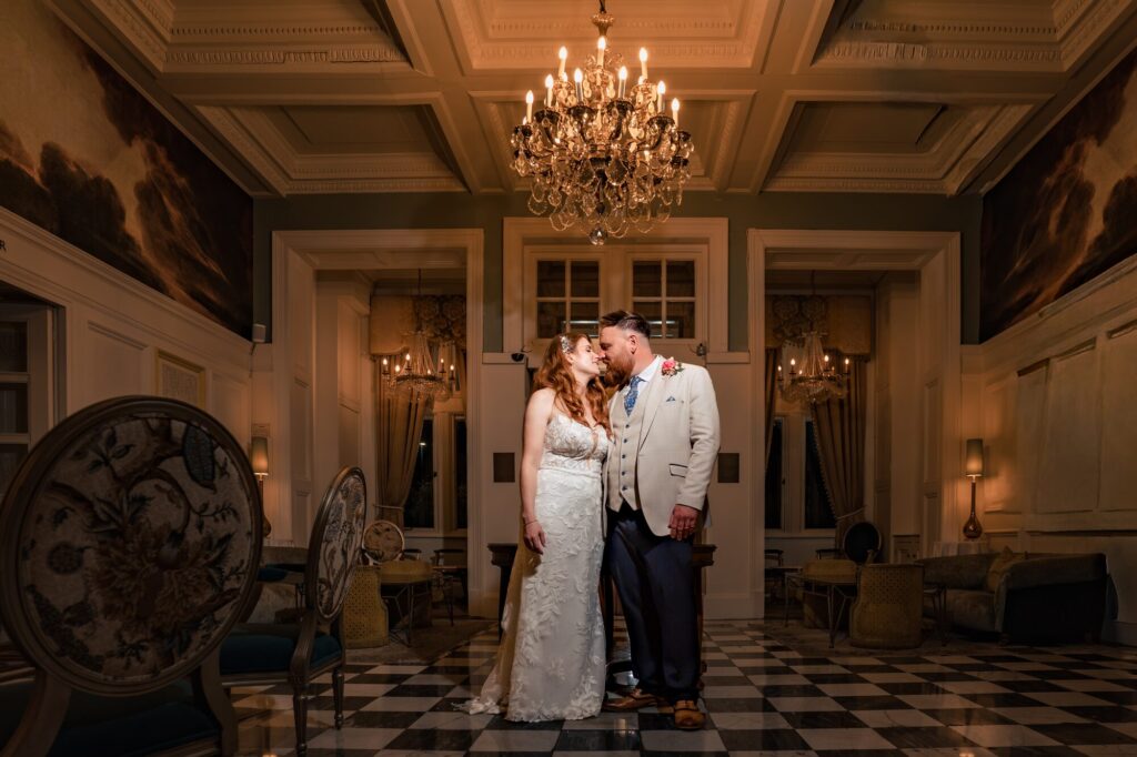 Bride and groom nuzzling noses beneath a crystal chandelier on the checkerboard floor at The Queens Hotel Southsea