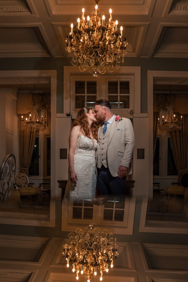 Bride and groom kissing beneath a crystal chandelier with a mirrored reflection at The Queens Hotel Southsea