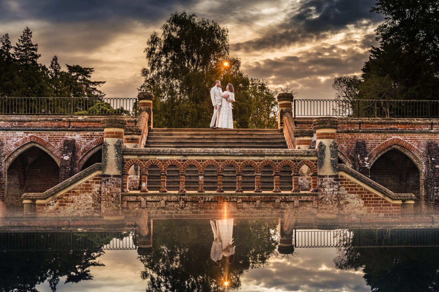 Rhys and Harpreet on the red brick staircase at Staunton Country Park with a mirror reflection and a flash-lit sunburst, creative post-wedding shoot
