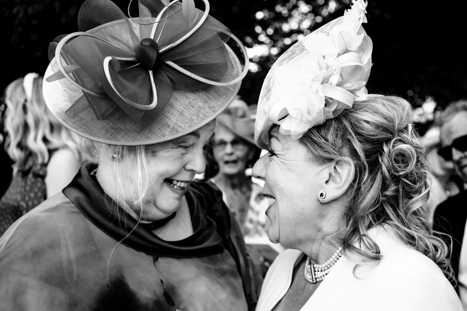 Two wedding guests in fascinators sharing a big laugh, black and white