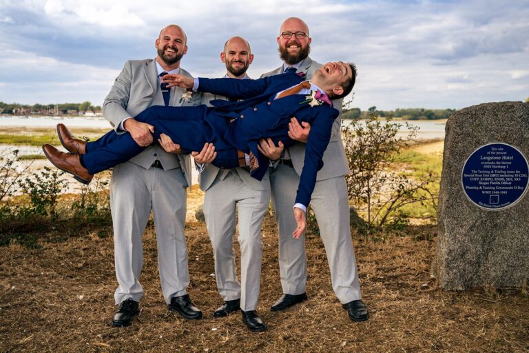 Groomsmen holding the groom horizontally like a plank for a fun group photo by the water