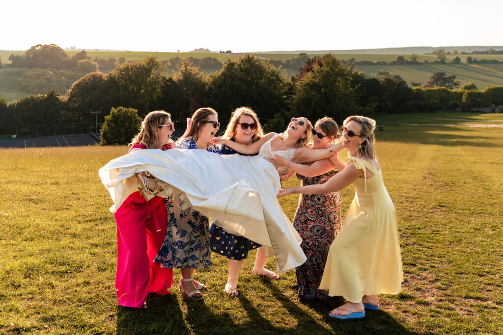 Bridesmaids carrying the bride across the field at golden hour at Great Ballard School, Eartham