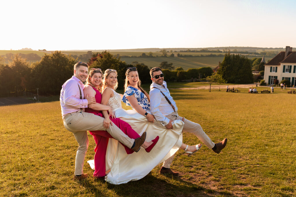 Bridesmaids and groomsmen lifting the bride in the evening sunshine at Great Ballard School, Eartham